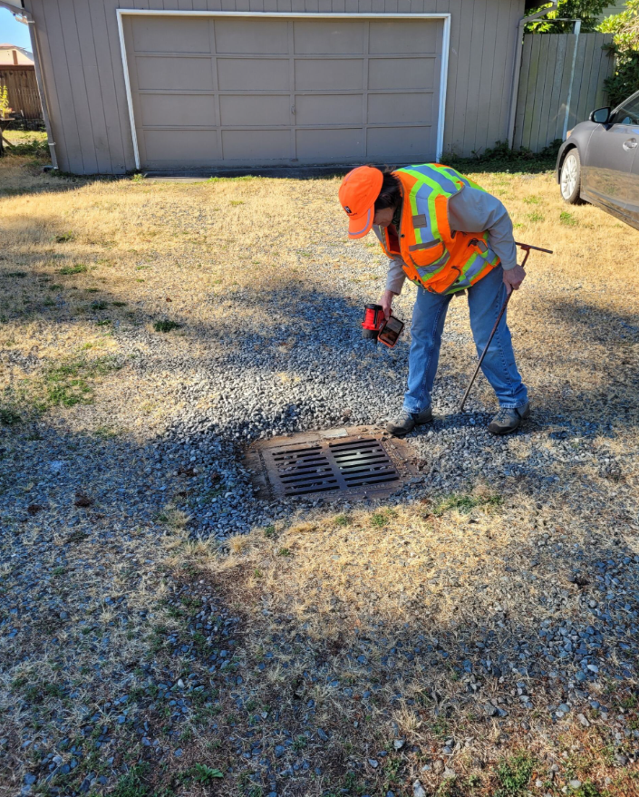 A person in an orange hat and vest looks into a storm drain