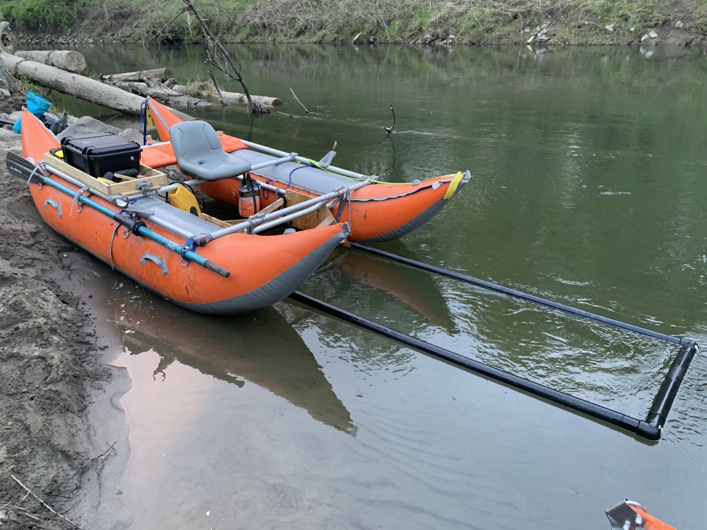 A gray seat is attached to two large orange pontoons at the edge of a calm river
