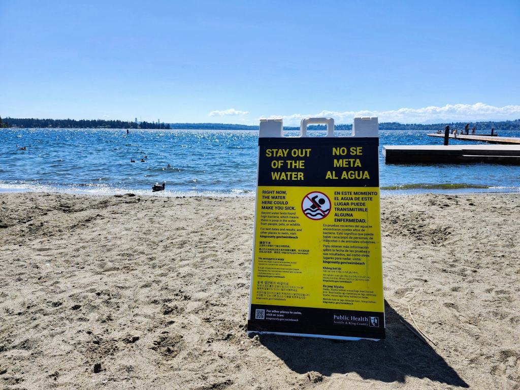 A large yellow and black sign on a sandy beach warns people to stay out of the water.