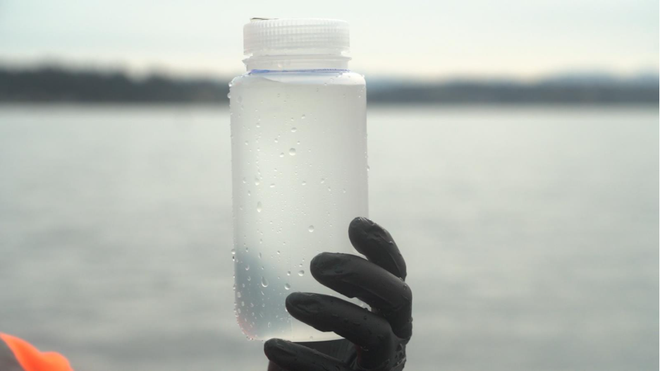 A gloved hand holds up a clear sample bottle full of water