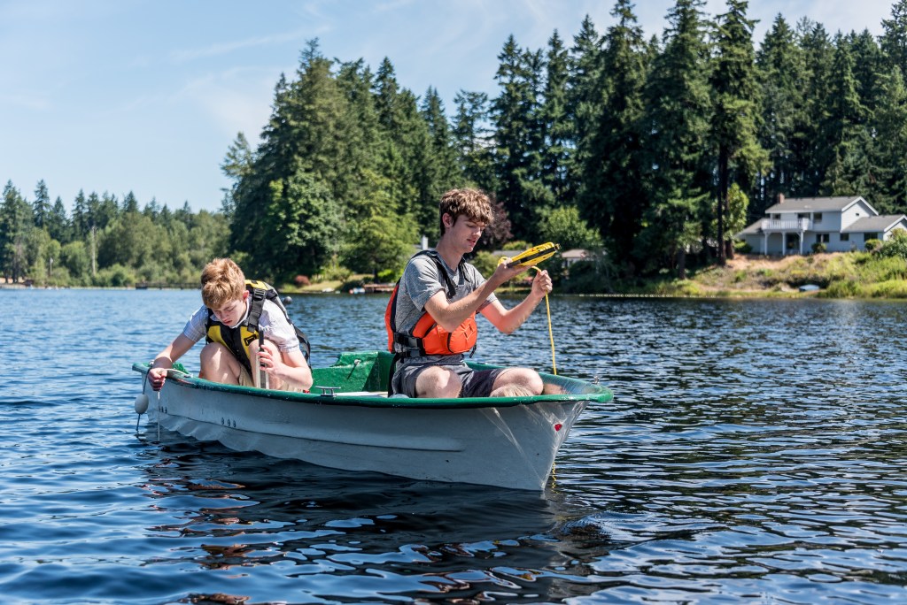 Two teenagers wearing life jackets sit in a boat with water sampling equipment