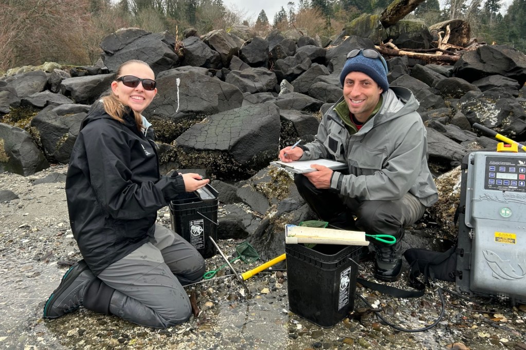 A person in a black jacket and sunglasses kneels on a wet rocky beach beside another person in a grey jacket. They are smiling, surrounded by buckets and fishing gear, in front of a pile of black rocks.