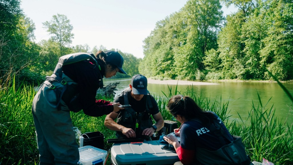 Three people in blue gear crouch together around measuring equipment. A river surrounded by green vegetation is in the background.